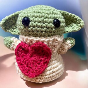 Puede incluir: Un juguete de peluche verde tejido a crochet con grandes ojos negros y un coraz&oacute;n rosa en el pecho. El juguete lleva un su&eacute;ter tejido a crochet de color crema.