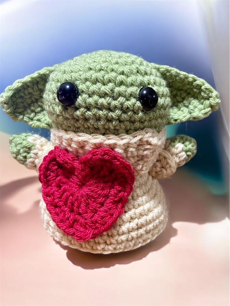 Puede incluir: Un juguete de peluche verde tejido a crochet con grandes ojos negros y un coraz&oacute;n rosa en el pecho. El juguete lleva un su&eacute;ter tejido a crochet de color crema.