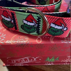 May include: A festive dog collar featuring a Grinch design with a red and white Santa hat on a green face, set against a red and green striped background. The collar has a black nylon base and a gold-colored metal D-ring.