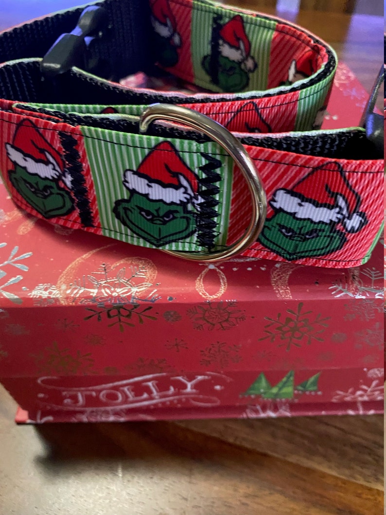 May include: A festive dog collar featuring a Grinch design with a red and white Santa hat on a green face, set against a red and green striped background. The collar has a black nylon base and a gold-colored metal D-ring.