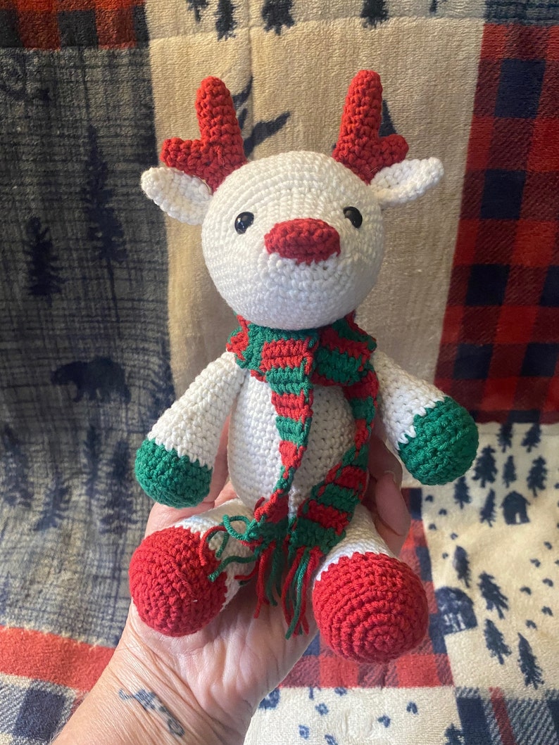 May include: White crocheted reindeer with red antlers, green and red striped scarf, and red crocheted feet.
