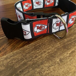 Puede incluir: Un collar de perro negro y rojo con un logotipo blanco y rojo de los Kansas City Chiefs repetido en todo el collar. El collar tiene una hebilla negra y un anillo en D plateado.