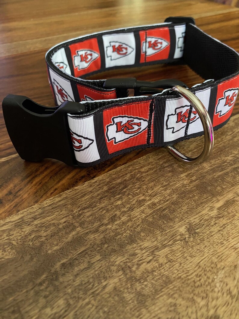 Puede incluir: Un collar de perro negro y rojo con un logotipo blanco y rojo de los Kansas City Chiefs repetido en todo el collar. El collar tiene una hebilla negra y un anillo en D plateado.