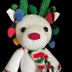 May include: A handmade white crochet reindeer with a red, white, and green scarf. The reindeer has green crochet antlers and is adorned with colorful Christmas lights. The reindeer has black eyes and a red, glittery nose.