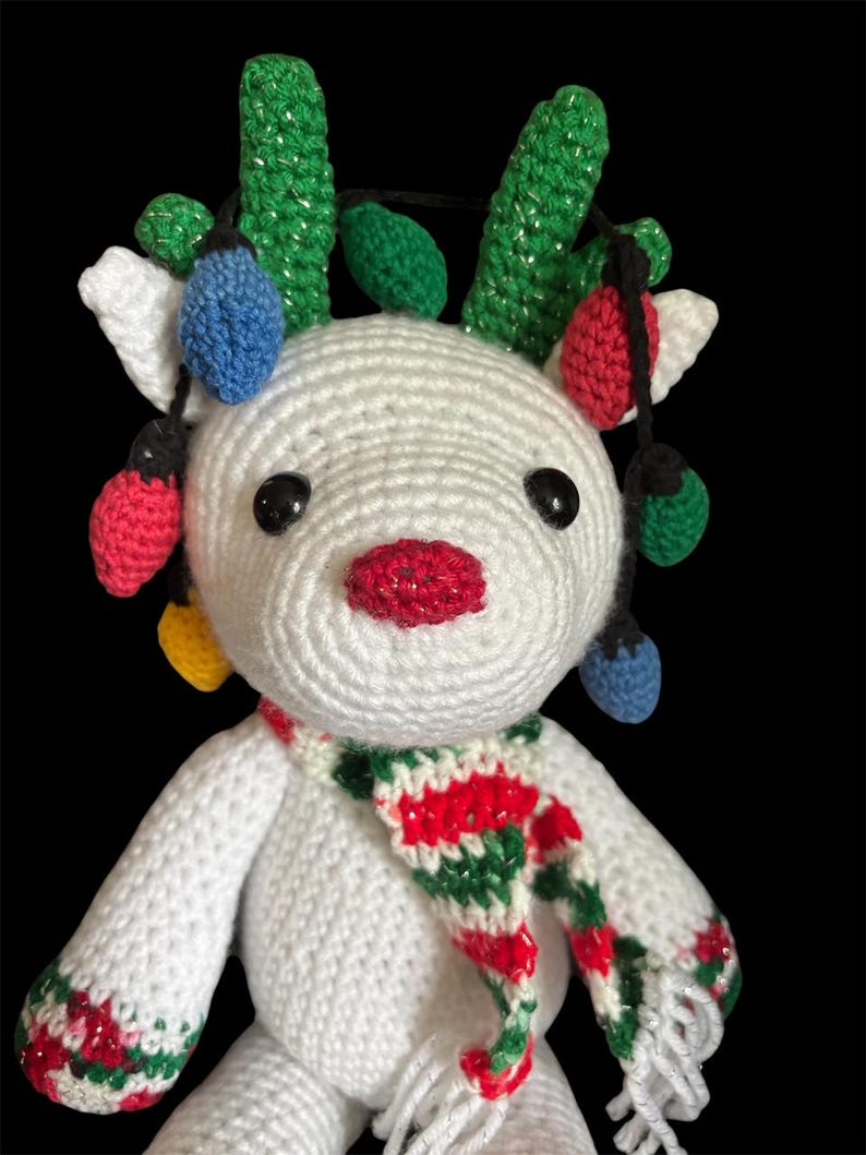 May include: A handmade white crochet reindeer with a red, white, and green scarf. The reindeer has green crochet antlers and is adorned with colorful Christmas lights. The reindeer has black eyes and a red, glittery nose.