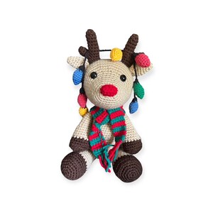 May include: A handmade crochet reindeer with a red nose, brown antlers, and a colorful scarf. The reindeer is adorned with a string of colorful Christmas lights. The plush toy is a festive holiday decoration.