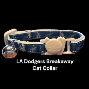 May include: A blue and white LA Dodgers breakaway cat collar with a white cat-shaped buckle. The collar has a silver bell and the words "LA Dodgers" printed on it. The text "LA Dodgers Breakaway Cat Collar" is below the collar.