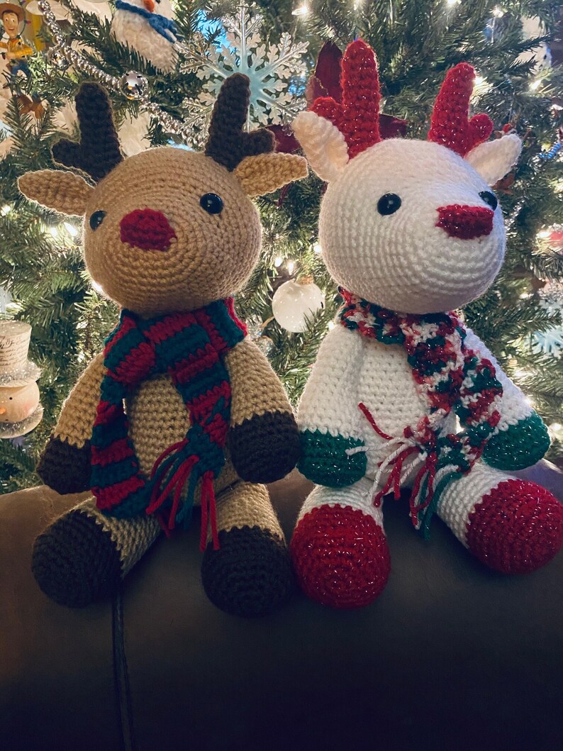 May include: Two handmade crochet reindeer dolls. One is tan with a red nose and a green, red, and blue scarf. The other is white with a red nose and a similar scarf. Both have dark brown antlers and hooves, and black eyes. Christmas decor.