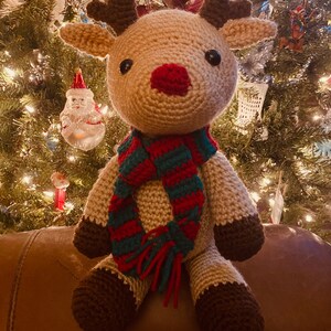 May include: A handmade crochet reindeer with a red nose, brown antlers, and a green and red striped scarf. The reindeer is sitting in front of a Christmas tree with lights and ornaments. The reindeer's hooves and the tips of its ears are brown.