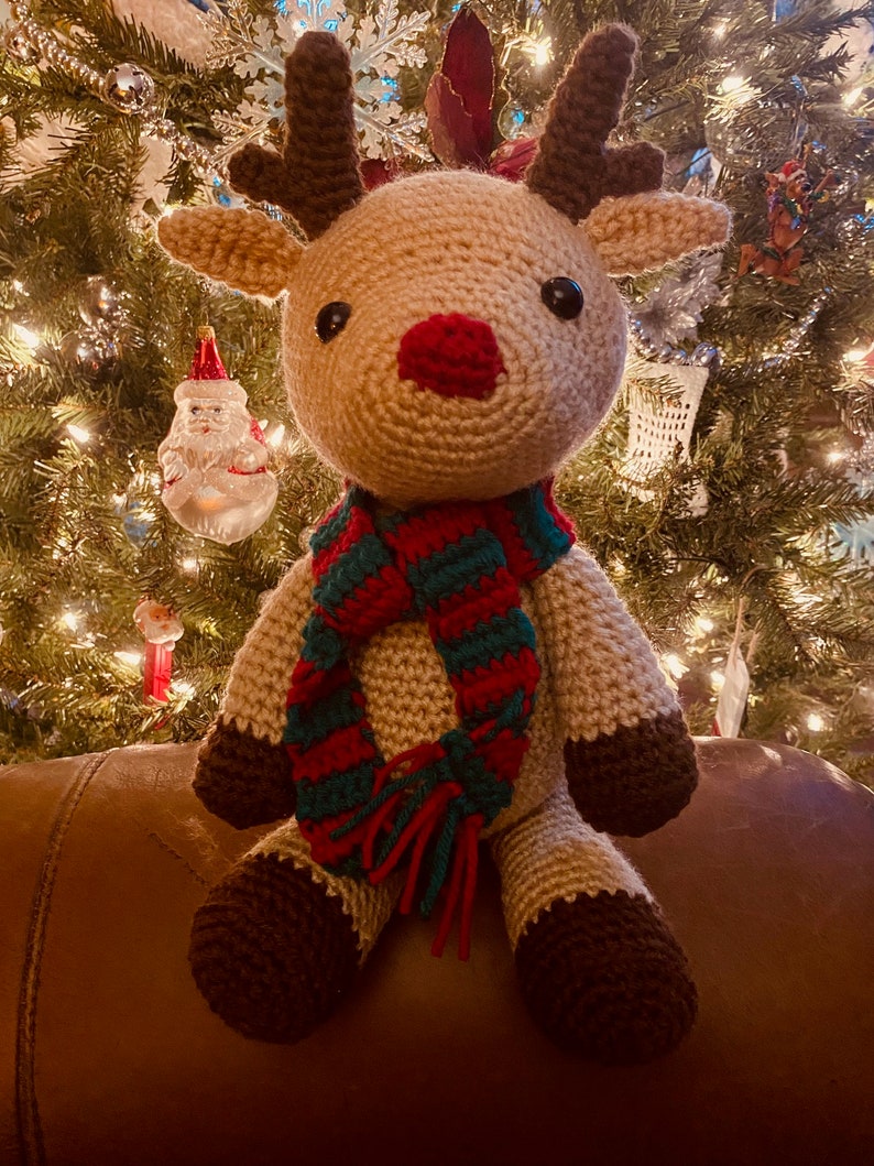 May include: A handmade crochet reindeer with a red nose, brown antlers, and a green and red striped scarf. The reindeer is sitting in front of a Christmas tree with lights and ornaments. The reindeer's hooves and the tips of its ears are brown.
