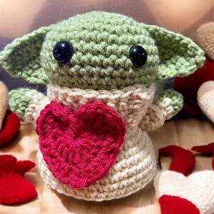Puede incluir: Un juguete de peluche verde tejido a crochet con grandes ojos negros y un coraz&oacute;n rosa en el pecho. El juguete lleva un su&eacute;ter blanco tejido a crochet.