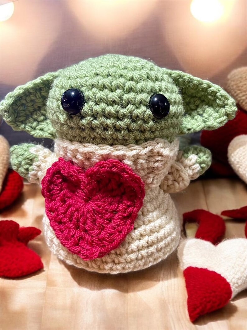 Puede incluir: Un juguete de peluche verde tejido a crochet con grandes ojos negros y un coraz&oacute;n rosa en el pecho. El juguete lleva un su&eacute;ter blanco tejido a crochet.