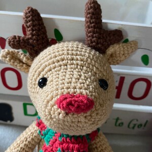 May include: A crocheted reindeer toy with brown antlers, a red nose, and a green and red scarf. The reindeer is sitting in front of a white wooden box with the words "HO HO" and "to Go!" written on it.