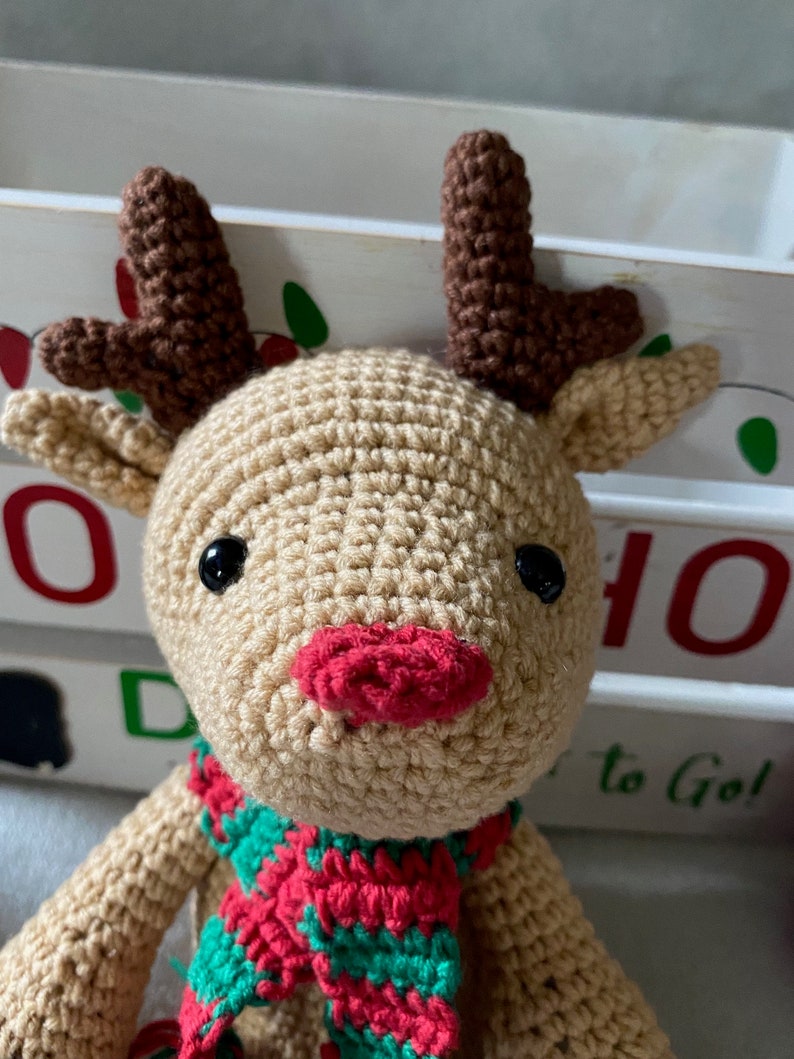 May include: A crocheted reindeer toy with brown antlers, a red nose, and a green and red scarf. The reindeer is sitting in front of a white wooden box with the words "HO HO" and "to Go!" written on it.