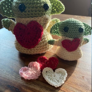 Puede incluir: Dos juguetes de peluche de crochet verde y beige con forma de un personaje ficticio popular, cada uno sosteniendo un coraz&oacute;n rojo. Cuatro corazones de crochet en rosa, rojo y blanco est&aacute;n sobre una superficie de madera.