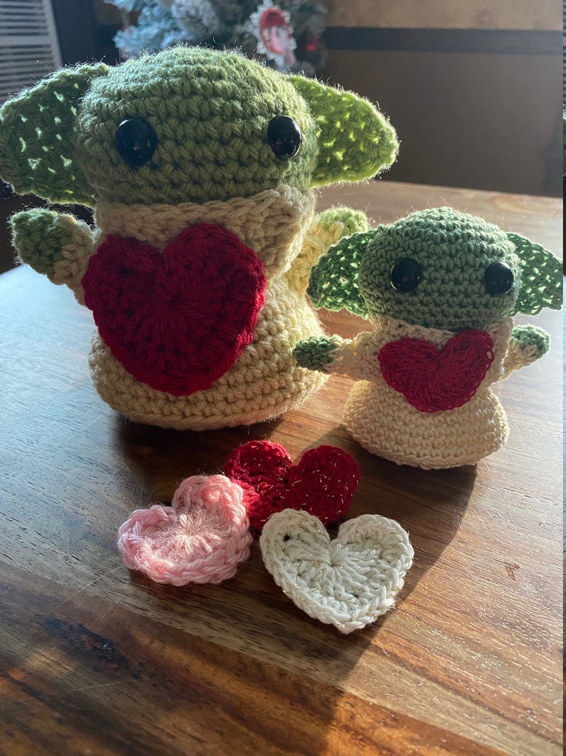 Puede incluir: Dos juguetes de peluche de crochet verde y beige con forma de un personaje ficticio popular, cada uno sosteniendo un coraz&oacute;n rojo. Cuatro corazones de crochet en rosa, rojo y blanco est&aacute;n sobre una superficie de madera.
