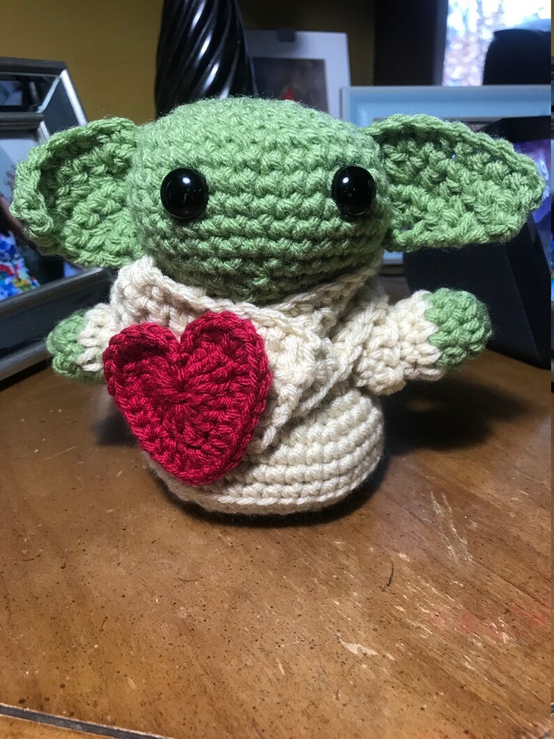 Puede incluir: Un juguete de peluche verde tejido a crochet con orejas grandes, ojos negros y un su&eacute;ter blanco tejido a crochet con un coraz&oacute;n rojo en la parte delantera.