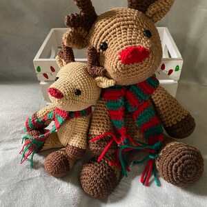May include: Two crocheted reindeer toys, one larger than the other, both wearing red and green striped scarves. The larger reindeer has brown antlers and a red nose. The smaller reindeer has brown antlers and a red nose.