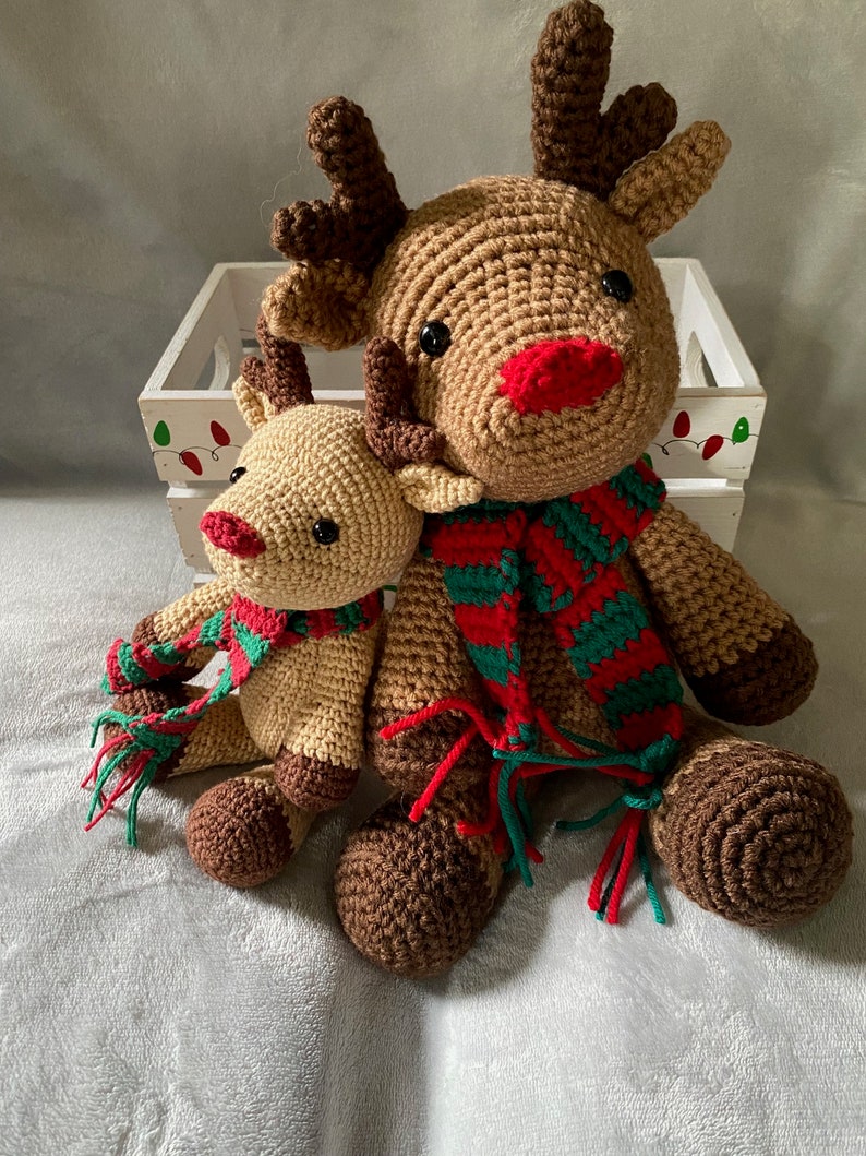 May include: Two crocheted reindeer toys, one larger than the other, both wearing red and green striped scarves. The larger reindeer has brown antlers and a red nose. The smaller reindeer has brown antlers and a red nose.