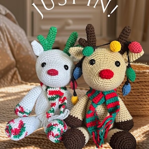 May include: Two crocheted reindeer dolls. One is white with green antlers and a red, green, and white scarf. The other is tan with brown antlers and a red, green, and white scarf. Both have Christmas lights and the text "JUST IN!" above them.