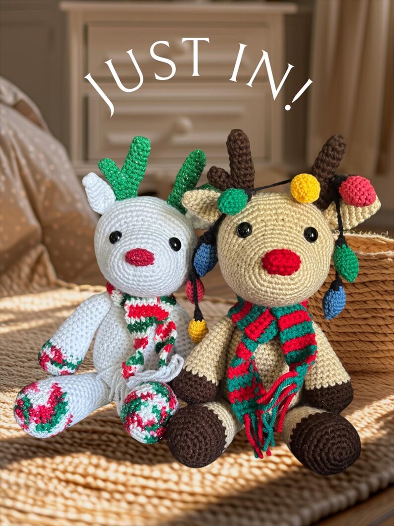 May include: Two crocheted reindeer dolls. One is white with green antlers and a red, green, and white scarf. The other is tan with brown antlers and a red, green, and white scarf. Both have Christmas lights and the text "JUST IN!" above them.