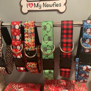 May include: A collection of dog collars with various patterns, including leopard print, snowmen, reindeer, and plaid. Each collar has a black buckle and a metal D-ring. The collars are displayed on a silver bar, with a sign that says "I My Newfies".
