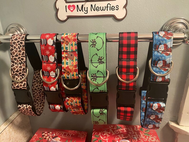 May include: A collection of dog collars with various patterns, including leopard print, snowmen, reindeer, and plaid. Each collar has a black buckle and a metal D-ring. The collars are displayed on a silver bar, with a sign that says "I My Newfies".