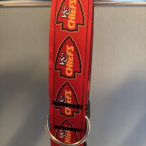 Puede incluir: Collar rojo para perro con un logotipo negro y amarillo de los Kansas City Chiefs repetido en el collar. El collar tiene un anillo en D de metal plateado.