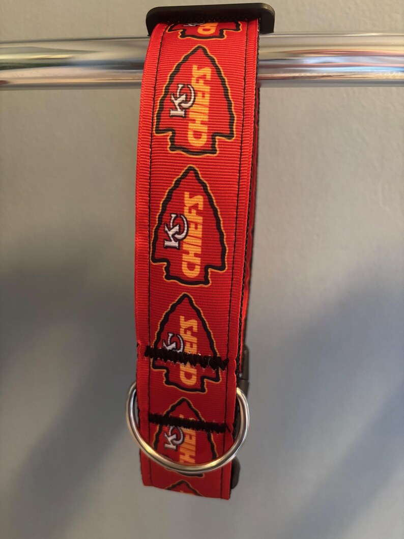 Puede incluir: Collar rojo para perro con un logotipo negro y amarillo de los Kansas City Chiefs repetido en el collar. El collar tiene un anillo en D de metal plateado.