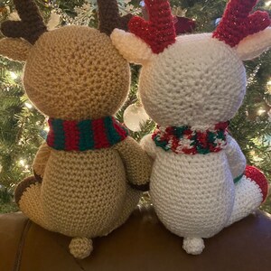 May include: Two handmade crochet reindeer dolls. One is tan with brown antlers and a red and green scarf. The other is white with red glitter antlers and a red, green, and white scarf. Both have small tails and are sitting.