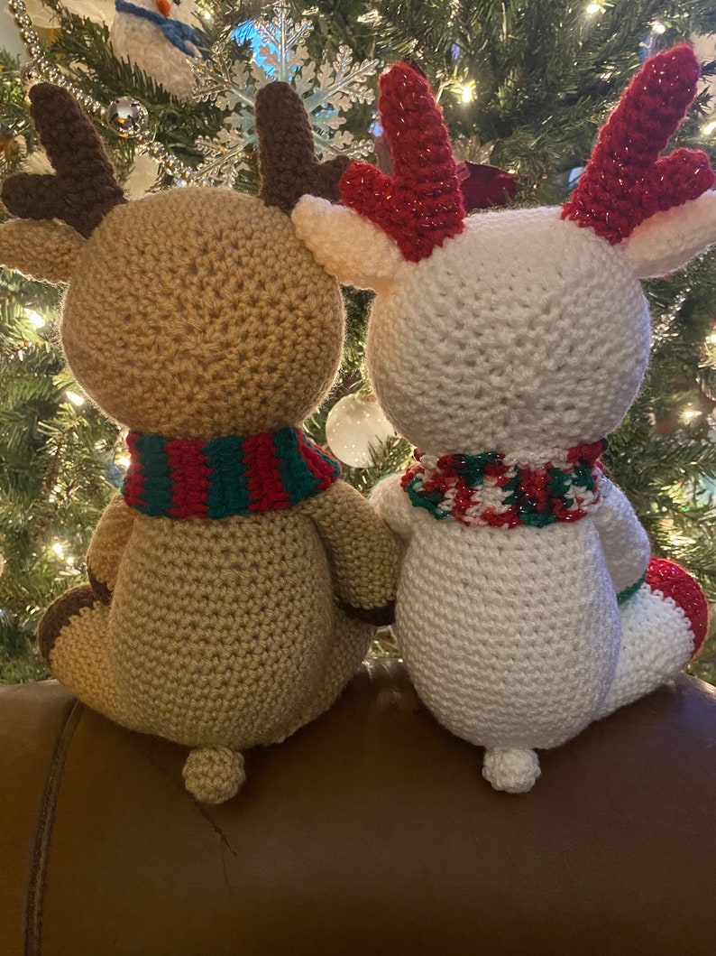 May include: Two handmade crochet reindeer dolls. One is tan with brown antlers and a red and green scarf. The other is white with red glitter antlers and a red, green, and white scarf. Both have small tails and are sitting.