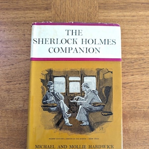 以下が含まれることがあります： 黄色い背景に、列車の車両に座っている2人の男性の白黒のイラストが描かれた本の表紙。本のタイトルは「The Sherlock Holmes Companion」、著者は「Michael and Mollie Hardwick」。