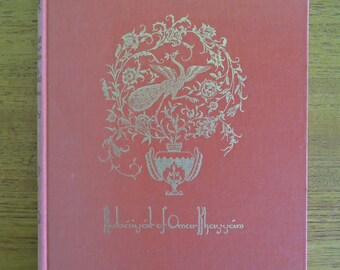 Könnte beinhalten: Ein gebundenes Buch mit lachsfarbenem Einband. Der Einband zeigt ein goldenes Blumendesign mit einem Vogel und dem Titel "Rubaiyat of Omar Khayyam" in goldener Schrift. Das Buch ist geschlossen und liegt auf einer Holzoberfläche.
