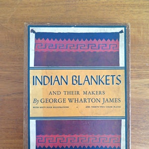 May include: A vintage book titled "Indian Blankets and Their Makers" by George Wharton James. The cover features a decorative border with geometric patterns in red, blue, and white. The book includes sixty-four illustrations and thirty-two color plates.