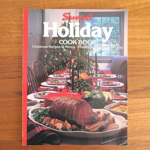 May include: A vintage "Sunset Holiday Cook Book" with a red spine. The cover features a festive holiday table setting with a roast, vegetables, and a decorated Christmas tree in the background. The book includes Christmas recipes and menus.