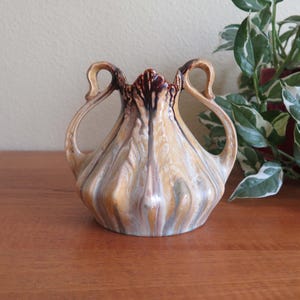 May include: A ceramic vase with a bulbous body and two arched handles. The vase features a blend of colors, including beige, brown, and blue, with a textured, marbled appearance. The top rim is a darker brown.