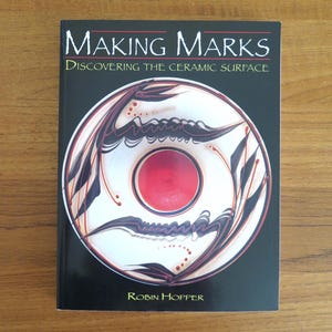 Puede incluir: Un libro titulado "MAKING MARKS Discovering the Ceramic Surface" de Robin Hopper. La portada es negra con texto blanco y un diseño circular con formas abstractas rojas, blancas y marrones. El libro está sobre una superficie de madera.