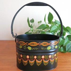 May include: A black metal oval-shaped bucket with a black handle. The bucket is painted with a colourful folk art design featuring yellow, red, orange, and green accents.