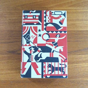 May include: A vintage book with a colorful circus-themed cover. The cover features geometric shapes and illustrations of circus performers in red, black, and white. The book is set against a wooden surface.