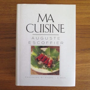 May include: A hardcover cookbook titled "MA CUISINE" by Auguste Escoffier. The cover features the title in black font, with a food image of rolled meat on a bed of greens. The book has a white cover and is on a wooden surface.