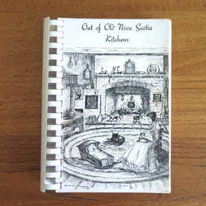 May include: A spiral-bound cookbook titled Out of Old Nova Scotia Kitchens. The white cover features a black and white ink illustration of a person sitting on a round rug near a fireplace, with a cradle and a cat nearby, all in a kitchen setting.
