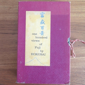 Puede incluir: Un libro rectangular magenta con una cubierta texturizada. Una etiqueta crema muestra texto japonés y las palabras en inglés "one hundred views of Fuji by Hokusai". Una cuerda está atada al costado del libro.