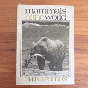May include: A book titled "mammals of the world" with a black and white image of a bear catching a fish. The book is a third edition and has a beige cover with black and gold lettering. The background is a wooden surface.