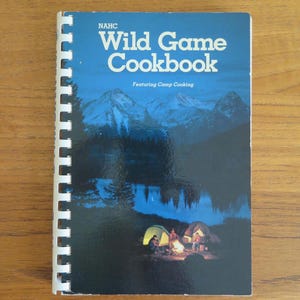 May include: A spiral-bound cookbook titled NAHC Wild Game Cookbook Featuring Camp Cooking. The cover features a blue-toned landscape illustration of mountains, a lake, and two tents with a campfire in the foreground at the bottom center.