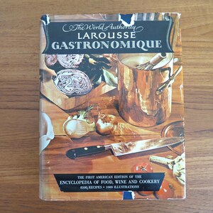 Puede incluir: Un libro de cocina vintage titulado "Larousse Gastronomique" con el texto "The World Authority" en la parte superior. La portada presenta una olla de cobre, un cuchillo, verduras y el texto "The First American Edition of the Encyclopedia of Food, Wine and Cookery".