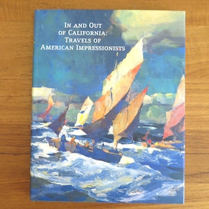 May include: A book cover with the title "In and Out of California: Travels of American Impressionists." The cover features a painting of sailboats on a choppy sea, with blue, orange, and white hues dominating the artwork. The book is set against a wooden surface.