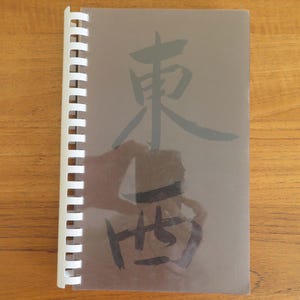 May include: A spiral-bound notebook with a translucent cover. The cover features black Japanese calligraphy. The notebook is open on a wooden surface. The text on the cover reads "東" and "西".