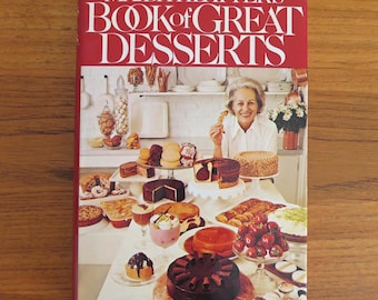 May include: A cookbook titled "Maida Heatter's Book of Great Desserts" with a red cover. The image features a variety of desserts, including cakes, pastries, and parfaits, arranged on a table. A woman is holding a pastry.