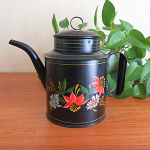May include: A black metal teapot with a floral design painted on the side. The flowers are red, yellow, and white. The teapot has a black handle and a black spout.
