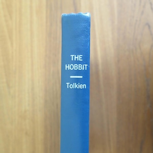 May include: A close-up of a blue book spine with the title "THE HOBBIT" and the author's name, Tolkien, in white lettering. The book's spine is a solid blue color, and the background is a light brown wooden surface.